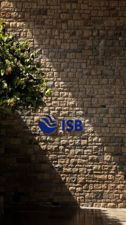 The blue ISB logo on a sunlit stone wall, with a tree in the corner